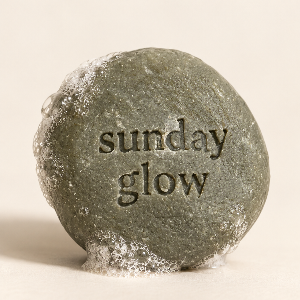 A round, textured gray stone with the words 'sunday glow' embossed on the front, set against a solid off-white background.